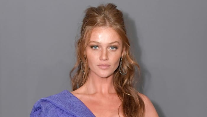 Cintia Dicker poses in a one-shoulder purple dress and wears her red hair in a half updo.